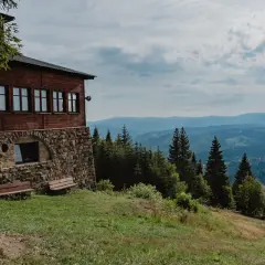 Drewniany budynek na zboczu góry z kamienną podmurówką, skąd rozpościera się widok na wielki stożek górski i lasy.