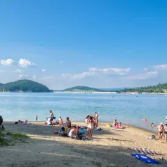 Słoneczna plaża w Polańczyku tętni życiem. Ludzie odpoczywają na piasku, kąpią się w jeziorze, a na horyzoncie widać żaglówkę.
