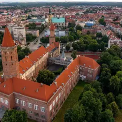 Legnica atrakcje: widok z lotu ptaka na zamek Piastowski z charakterystyczną wieżą i czerwonym dachem, otoczony zielenią i zabudową miejską.