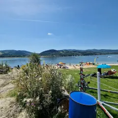 Słoneczna plaża nad Jeziorem Mucharskim tętni życiem. Widać ludzi odpoczywających na piasku, żaglówki na wodzie i zielone wzgórza w tle.
