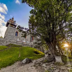 Kamienna wieża na górze Ślęża, otoczona zielenią i starym drzewem, z widokiem na zachodzące słońce.