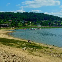 Słoneczna plaża nad Jeziorem Rożnowskim, z łódkami i domkami na wzgórzu.