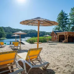 Piękny dzień na plaży nad jeziorem. Leżaki i parasole zapraszają do odpoczynku. W tle widać drewnianą saunę i zalesione wzgórza pojezierza brodnickiego.