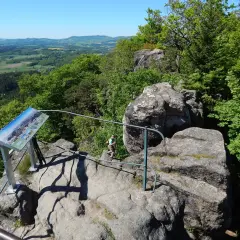 Widok z Krzyżnej Góry na zielone wzgórza. Turystka podziwia panoramę, obok tablica informacyjna.