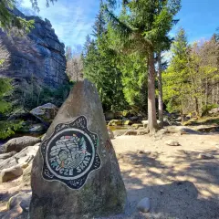 Kamień z logo Krucze Skały przy wejściu na via ferrata. W tle skalne ściany i las.