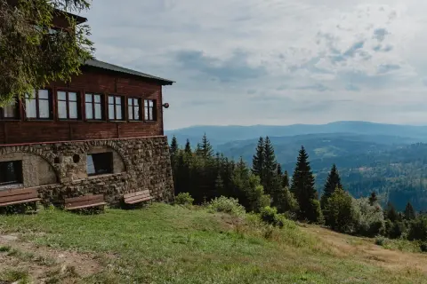 Drewniany budynek na zboczu góry z kamienną podmurówką, skąd rozpościera się widok na wielki stożek górski i lasy.