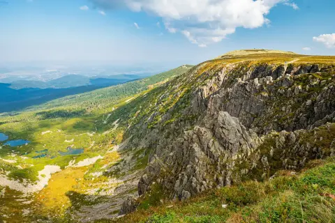 Widok na skaliste zbocze i zieloną dolinę z jeziorami. W oddali widać miasto. To krajobraz przypominający **śnieżne kotły**.