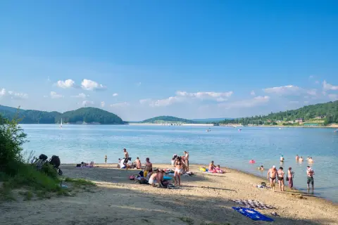 Słoneczna plaża w Polańczyku tętni życiem. Ludzie odpoczywają na piasku, kąpią się w jeziorze, a na horyzoncie widać żaglówkę.