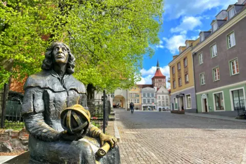 Pomnik Kopernika na Rynku Staromiejskim. Olsztyn co warto zobaczyć: urokliwe kamieniczki, zielone drzewa i błękitne niebo.