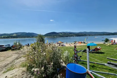 Słoneczna plaża nad Jeziorem Mucharskim tętni życiem. Widać ludzi odpoczywających na piasku, żaglówki na wodzie i zielone wzgórza w tle.