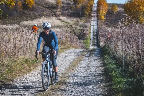 Kobieta na rowerze pokonuje kamienistą drogę w jesiennych Dolinkach Krakowskich. W tle widać innego rowerzystę.