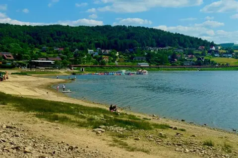 Słoneczna plaża nad Jeziorem Rożnowskim, z łódkami i domkami na wzgórzu.