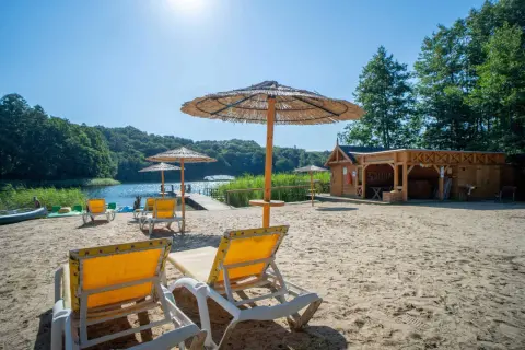 Piękny dzień na plaży nad jeziorem. Leżaki i parasole zapraszają do odpoczynku. W tle widać drewnianą saunę i zalesione wzgórza pojezierza brodnickiego.