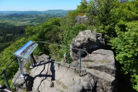 Widok z Krzyżnej Góry na zielone wzgórza. Turystka podziwia panoramę, obok tablica informacyjna.