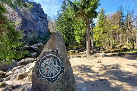 Kamień z logo Krucze Skały przy wejściu na via ferrata. W tle skalne ściany i las.