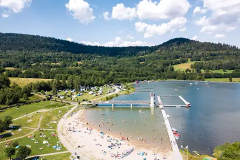 Słoneczny dzień nad zalewem Stara Morawa. Ludzie odpoczywają na plaży i w wodzie, a w tle widać zielone wzgórza.