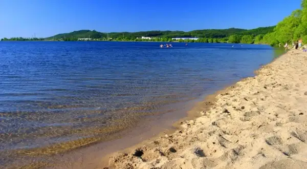 Słoneczny dzień nad Zalewem Sulejowskim. Piaszczysta plaża, błękitna woda i zielone wzgórza w tle. Idealne miejsce na relaks i wodne atrakcje.