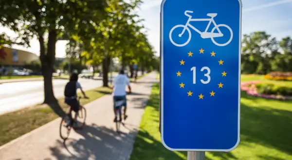 Znak EuroVelo 13 na ścieżce rowerowej. Dwie osoby jadą na rowerach wzdłuż alei drzew.
