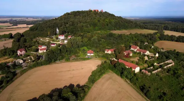 Krajobraz z ruinami zamku na wzgórzu, przypominający wygasłe wulkany w Polsce. Widać zabudowania i pola uprawne.