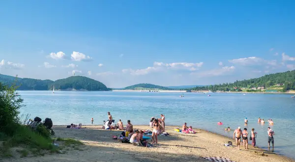 Słoneczna plaża w Polańczyku tętni życiem. Ludzie odpoczywają na piasku, kąpią się w jeziorze, a na horyzoncie widać żaglówkę.
