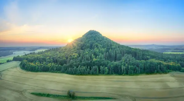 Wygasłe wulkany porośnięte lasem, otoczone polami zbóż, oświetlone wschodzącym słońcem.