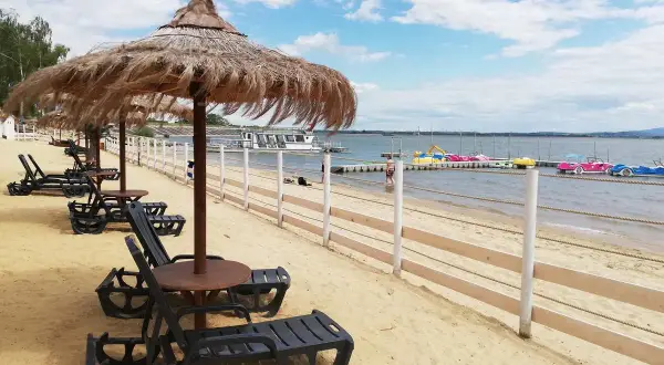Plaża nad Jeziorem Nyskim z leżakami i parasolami. W oddali widać wodne atrakcje: łódki i rowery wodne.