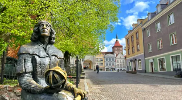 Pomnik Kopernika na Rynku Staromiejskim. Olsztyn co warto zobaczyć: urokliwe kamieniczki, zielone drzewa i błękitne niebo.