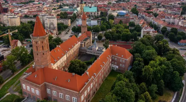 Legnica atrakcje: widok z lotu ptaka na zamek Piastowski z charakterystyczną wieżą i czerwonym dachem, otoczony zielenią i zabudową miejską.