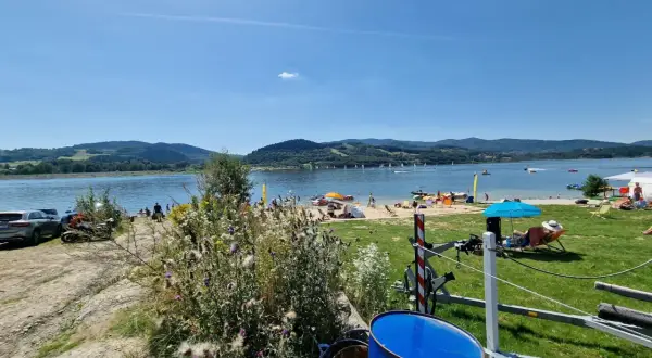 Słoneczna plaża nad Jeziorem Mucharskim tętni życiem. Widać ludzi odpoczywających na piasku, żaglówki na wodzie i zielone wzgórza w tle.