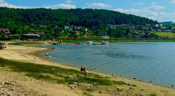 Słoneczna plaża nad Jeziorem Rożnowskim, z łódkami i domkami na wzgórzu.