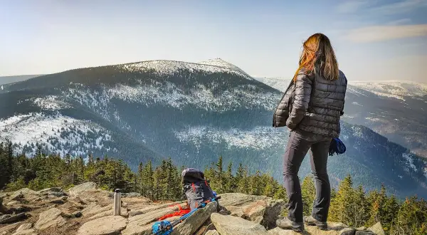 Kobieta na skalnym stole podziwia zimowy krajobraz gór. Plecak i kijki trekkingowe czekają na dalszą wędrówkę.
