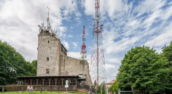 Kamienna wieża i maszty telekomunikacyjne na **chełmiec szczyt**. Ludzie odpoczywają na trawie pod drzewami.
