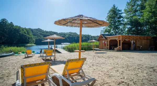 Piękny dzień na plaży nad jeziorem. Leżaki i parasole zapraszają do odpoczynku. W tle widać drewnianą saunę i zalesione wzgórza pojezierza brodnickiego.