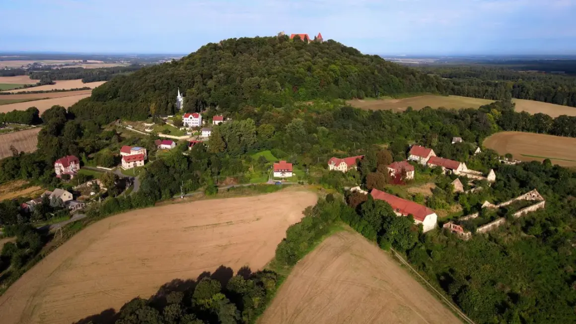 Krajobraz z ruinami zamku na wzgórzu, przypominający wygasłe wulkany w Polsce. Widać zabudowania i pola uprawne.