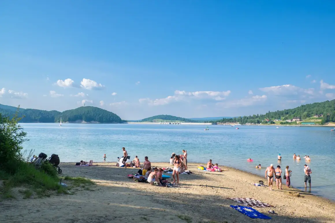 Słoneczna plaża w Polańczyku tętni życiem. Ludzie odpoczywają na piasku, kąpią się w jeziorze, a na horyzoncie widać żaglówkę.