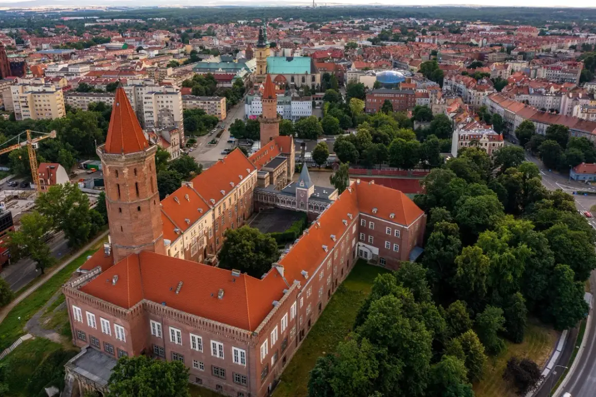 Legnica atrakcje: widok z lotu ptaka na zamek Piastowski z charakterystyczną wieżą i czerwonym dachem, otoczony zielenią i zabudową miejską.