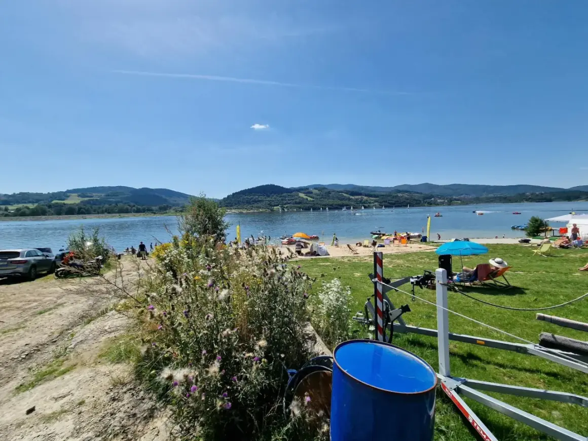 Słoneczna plaża nad Jeziorem Mucharskim tętni życiem. Widać ludzi odpoczywających na piasku, żaglówki na wodzie i zielone wzgórza w tle.