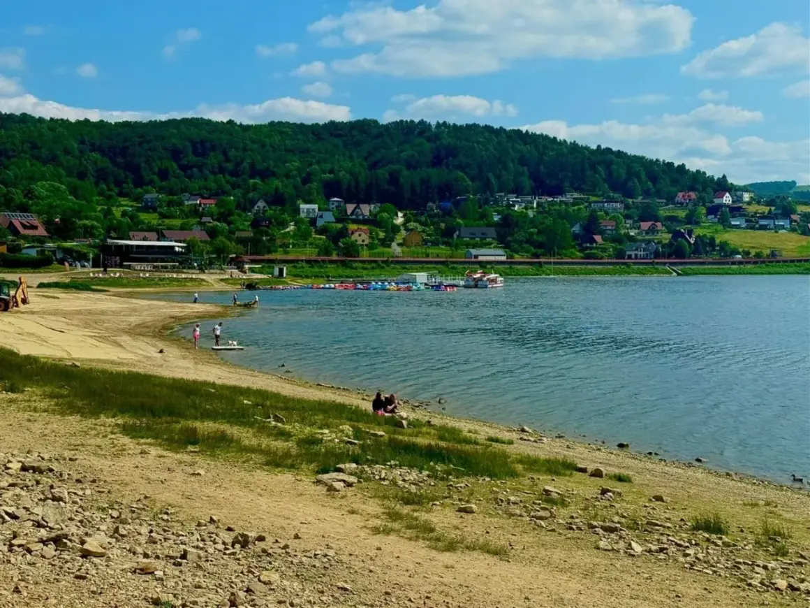 Słoneczna plaża nad Jeziorem Rożnowskim, z łódkami i domkami na wzgórzu.