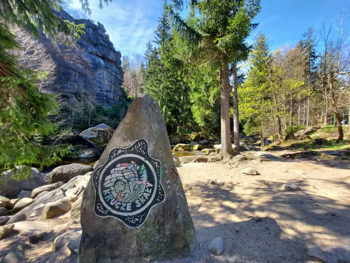 Kamień z logo Krucze Skały przy wejściu na via ferrata. W tle skalne ściany i las.