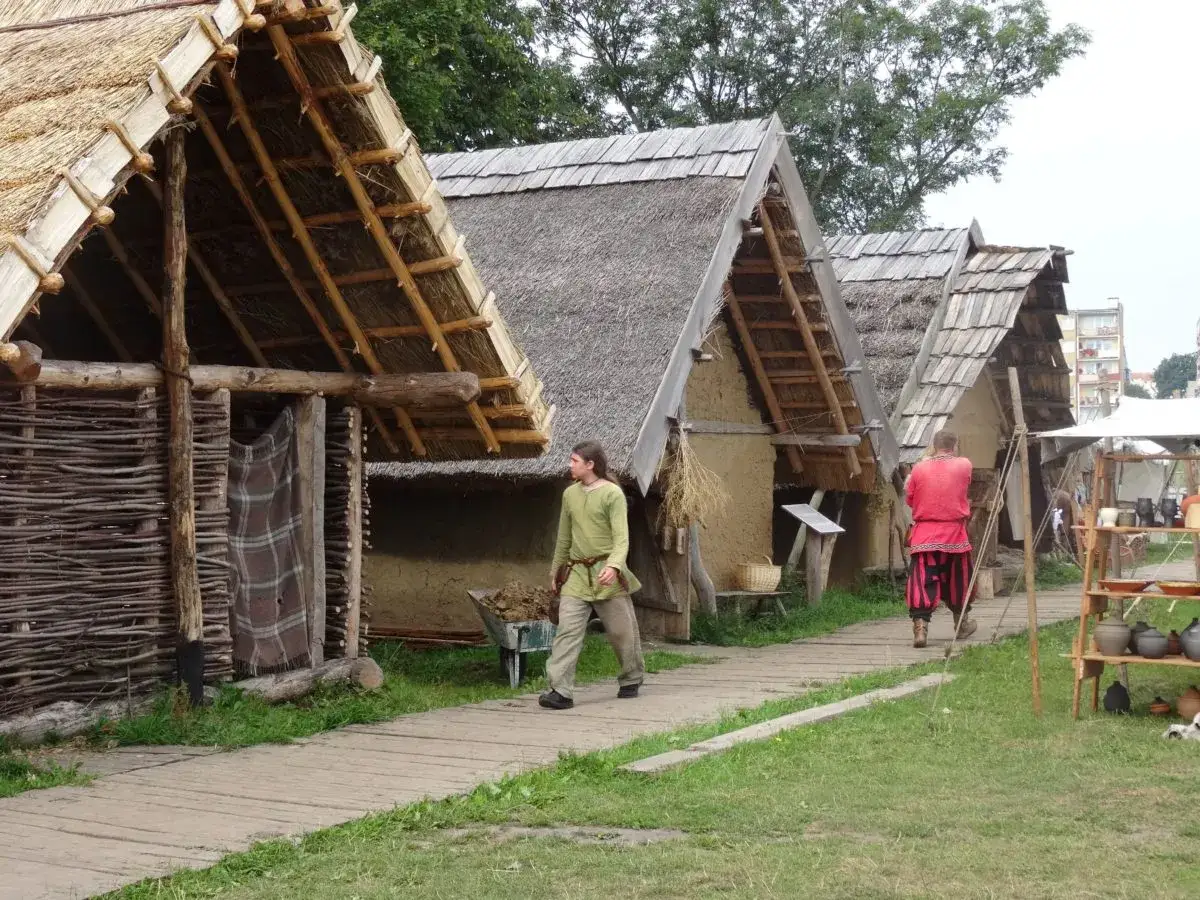 Wioska wikingów Wolin. Drewniane chaty kryte strzechą, mężczyźni w historycznych strojach spacerują po drewnianej ścieżce.