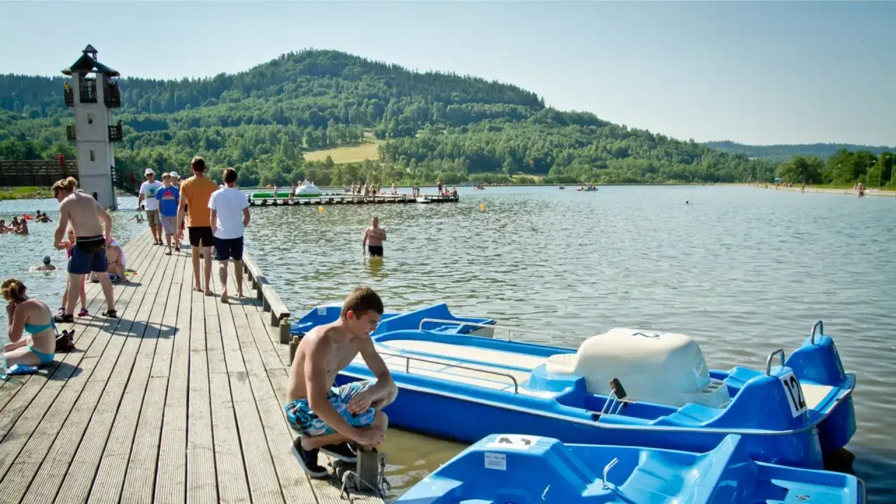 Letnia rekreacja nad jeziorem, gdzie można podziwiać śląskie atrakcje. Ludzie odpoczywają na pomoście, pływają i korzystają z rowerów wodnych.