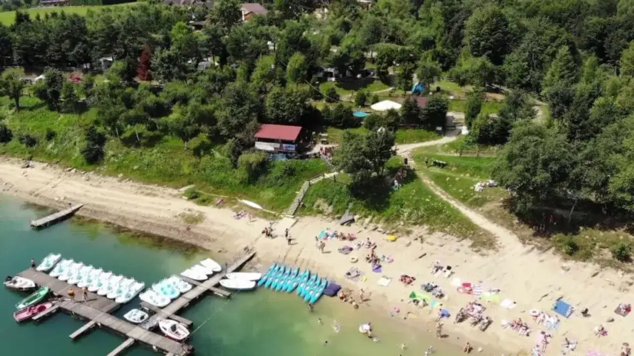 Plaża w Polańczyku tętni życiem. Widać ludzi odpoczywających na piasku, pływających w jeziorze i korzystających z wypożyczonych łódek oraz rowerków wodnych.