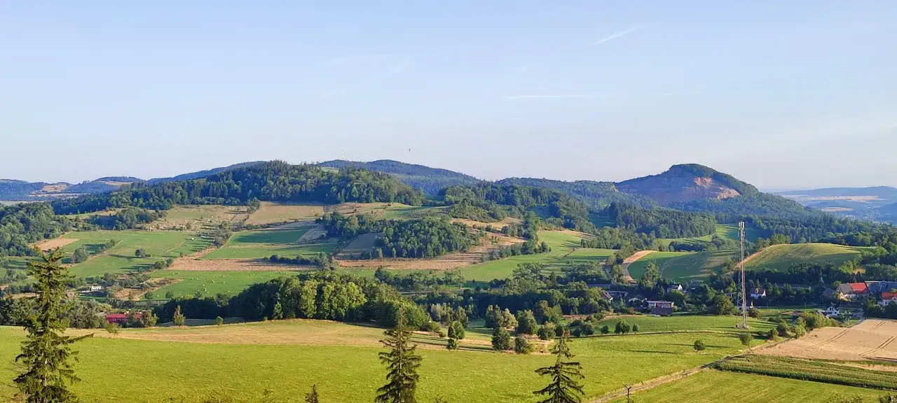 Zielone wzg&oacute;rza i pola tworzą malowniczy krajobraz, przypominający krainę wygasłych wulkan&oacute;w. W oddali widać lasy i niewielką wioskę.