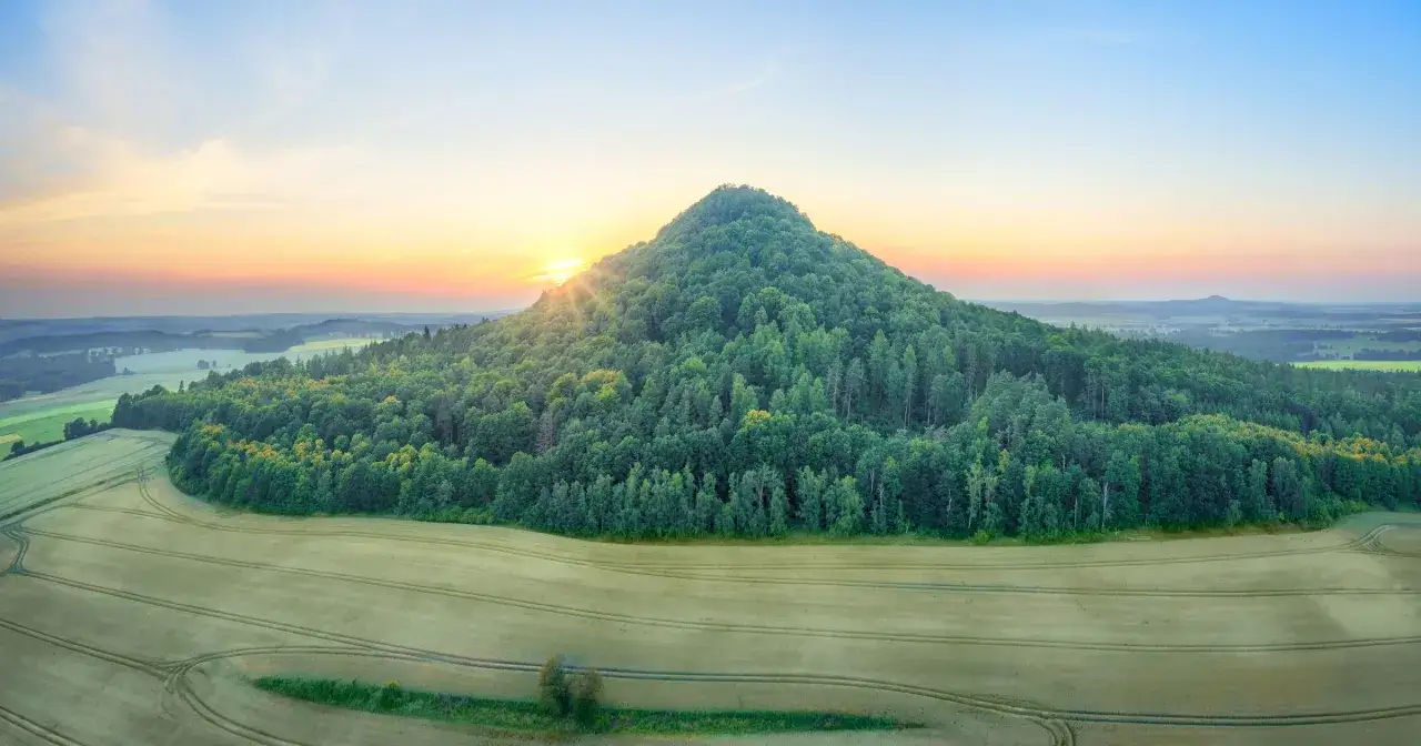 Wygasłe wulkany porośnięte lasem, otoczone polami, w blasku wschodzącego słońca.