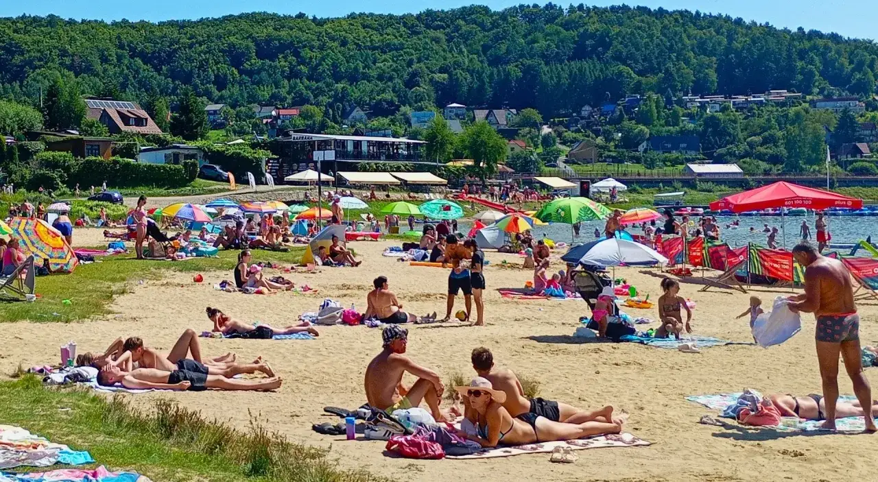 Słoneczna plaża nad Jeziorem Rożnowskim tętni życiem. Ludzie odpoczywają pod parasolami, kąpią się i opalają.