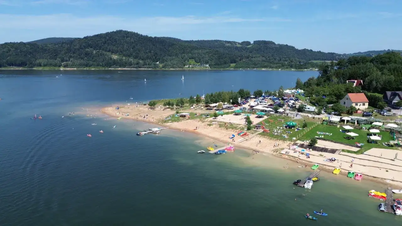 Słoneczna plaża nad Jeziorem Mucharskim tętni życiem. Ludzie opalają się, pływają i relaksują na piasku.