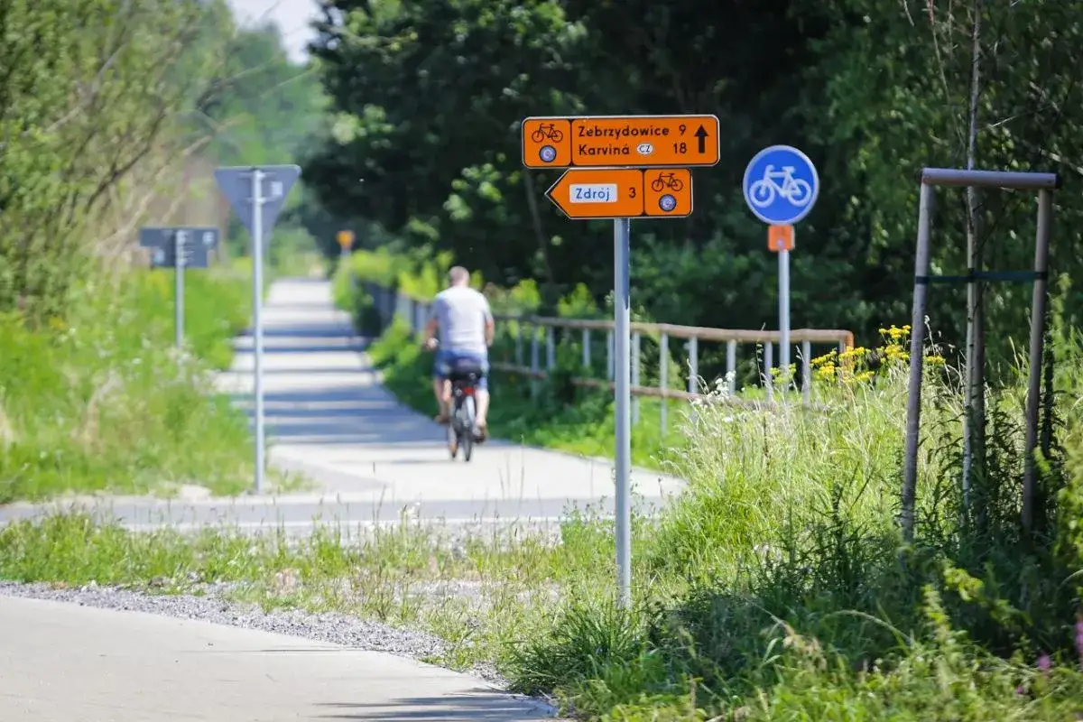 Rowerzysta jedzie ścieżką, mijając znaki wskazujące żelazny szlak rowerowy do Zebrzydowic i Karwiny.