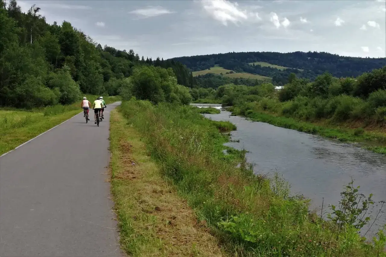 Trzy osoby na rowerach jadą ścieżką rowerową wzdłuż rzeki. Malowniczy krajobraz Beskidu Sądeckiego zachęca do aktywnego wypoczynku, idealnego na velo krynica.