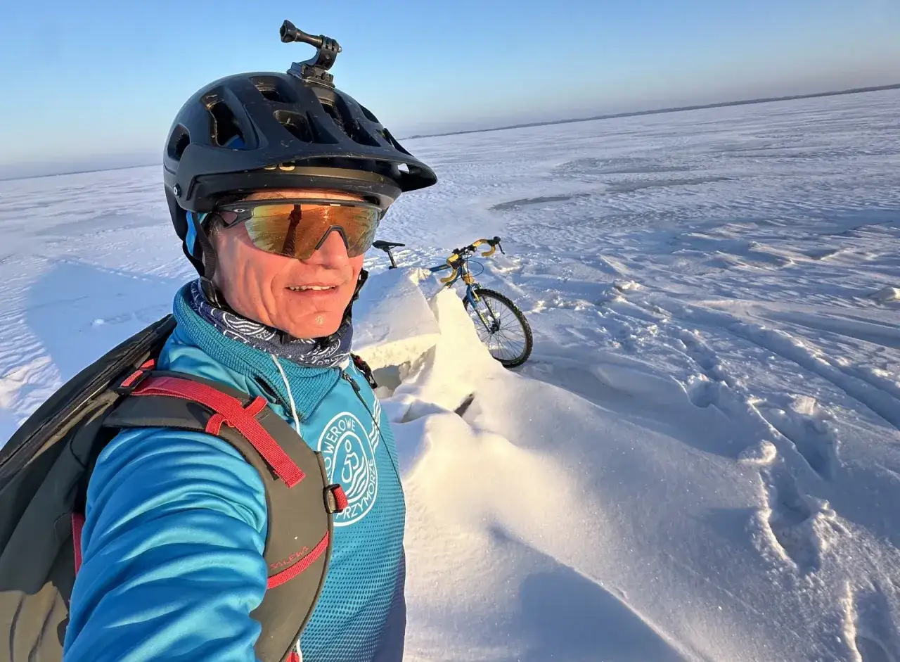 Rowerzysta w kasku z kamerą GoPro na głowie pozuje na tle zamarzniętego jeziora. W tle rower, idealny na zimowe trasy rowerowe zachodniopomorskie.
