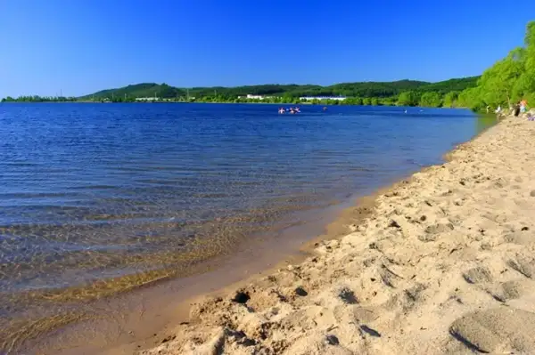 Słoneczny dzień nad Zalewem Sulejowskim. Piaszczysta plaża, błękitna woda i zielone wzgórza w tle. Idealne miejsce na relaks i wodne atrakcje.